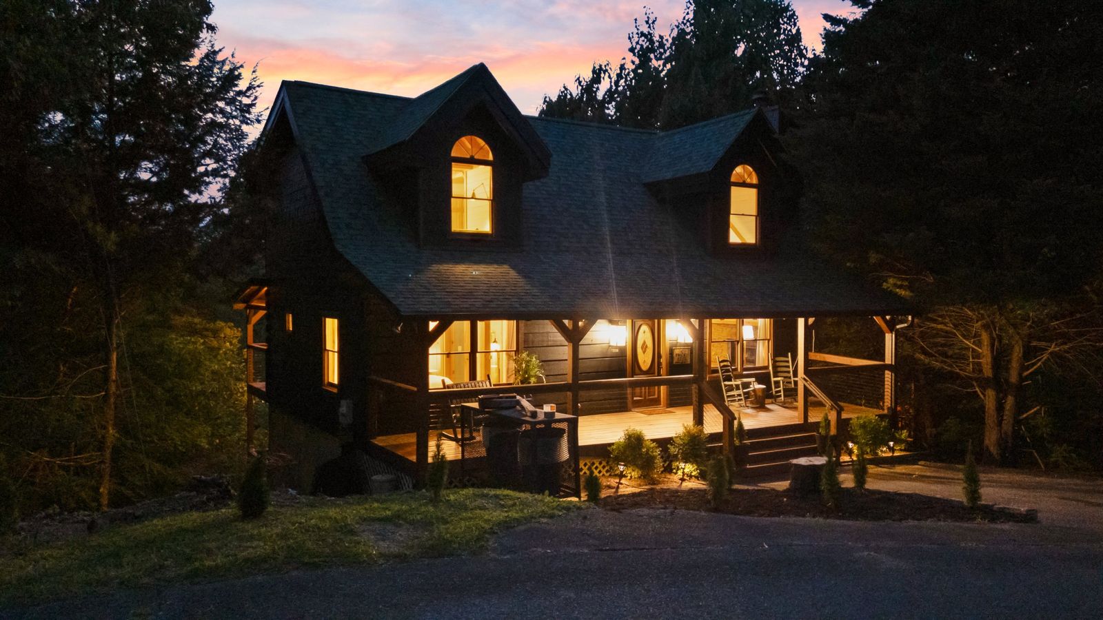 The Smoky Edit log cabin at dusk, porch and dormer windows glowing beneath a pink sky